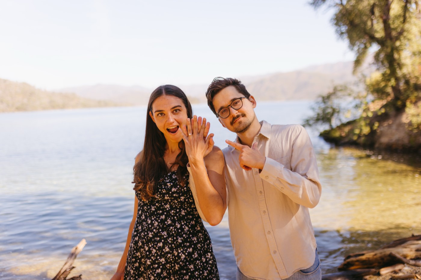 Andrea showing her ring to the camera while Alex points at it, the lake behind them