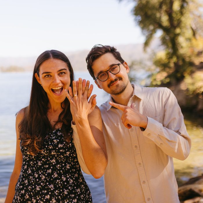 Andrea holding up her hand to show the ring, Alex pointing at it, both laughing beside the lake